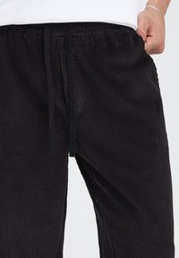 Pantaloni negri din catifea cu dungi, cu textură nervurată, talie elastică cu șnur și buzunare laterale, văzuți de la talie în jos.