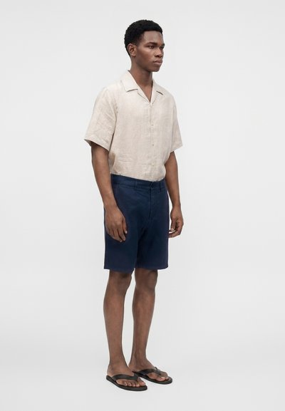 Homme portant une chemise beige à manches courtes boutonnée, un short bleu marine et des tongs noires, debout devant un fond blanc uni.