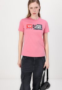Camiseta de manga corta rosa con un logo gráfico en blanco y negro, combinada con pantalones cargo gris oscuro. Textura de algodón suave. Bolso negro.