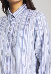 Camicia in lino a righe blu chiaro e bianche con colletto, chiusura frontale con bottoni e taschino sul petto. Tessuto leggermente stropicciato.