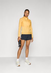 Veste légère jaune avec fermeture éclair à l'avant, coupe ajustée et capuche. Associée à un short sombre. Le sujet tient une bouteille d'eau noire.