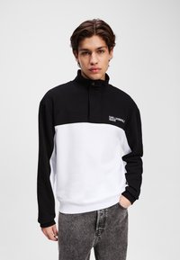Pullover in bianco e nero con col alto, dotato di un logo sul petto, maniche lunghe e un tessuto morbido e testurizzato.