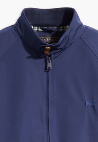 Marineblaue, leichte Jacke mit hohem Kragen, Reißverschluss, zwei braunen Knöpfen und karierter Innenfüllung. Mit kleinem Logo auf der Brust.