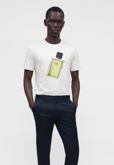 Homme portant un t-shirt blanc avec un grand motif de flacon de parfum et un pantalon bleu foncé, debout devant un fond clair uni.