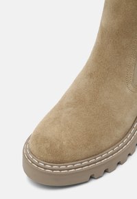 Beige Wildleder-Absatzschuh mit runder Zehe, ausgestattet mit einer dicken, strukturierten Gummisohle und kontrastierenden weißen Nähten entlang des Randes.