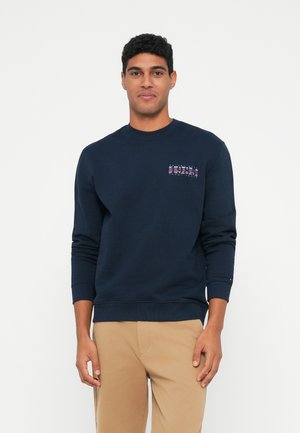 Mies, der einen navyblauen Sweatshirt mit "1985"-Logo auf der linken Brust und beigen Hosen trägt, steht vor einem weißen Hintergrund.
