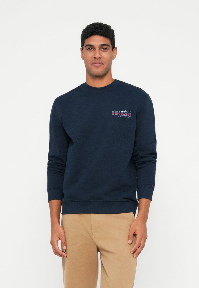 Moški v temnoplavem jopiču s logotipom "1985" na levem prsnem delu in bež hlačah, stoji pred belo ozadjem.