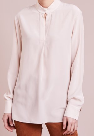 Femme portant un chemisier rose pâle à manches longues avec un col mandarin et des poignets boutonnés, associé à un pantalon marron.
