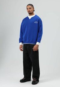 Blauwe trui met witte kraagdetails, losse pasvorm, gecombineerd met zwarte wijde jeans en zwarte schoenen met veteraccenten.