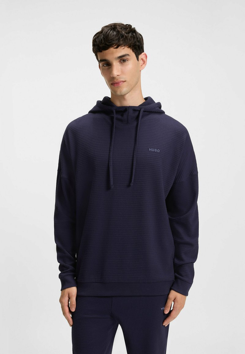 Granatowa bluza z kapturem o teksturowanym wykończeniu, ze sznurkami przy szyi i subtelnym logo na klatce piersiowej, z luźnym krojem i długimi rękawami.