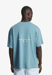 Prevelik svetlo modri bombažni T-shirt s kratkimi rokavi, ki ima na hrbtni strani beli tiskani napis in grafični dizajn.