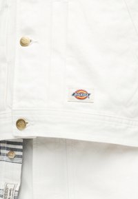 Λευκό μπουφάν Dickies με χρυσά κουμπιά και μοτίβο λογότυπου κεντημένο, φορεμένο πάνω από ριγέ πουκάμισο με μπεζ κουμπί.