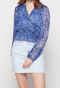 Blå blomsterinspirerad wrap-blus med genomskinliga långa ärmar och smockad midja, matchad med en ljusblå jeansminikjol.