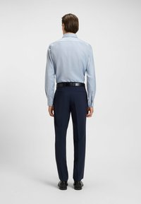 Lichtblauwe blouse met lange mouwen en een subtiel patroon, gecombineerd met donkerblauwe trousers. Een zwarte riem en schoenen maken de outfit af.