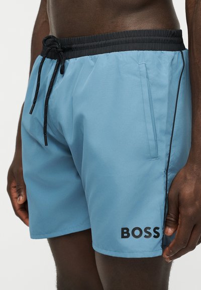 Short de bain bleu clair avec taille élastique noire et cordon, poches latérales, et logo noir "BOSS" sur la jambe gauche porté par une personne.