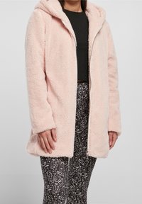 Chaqueta de forro polar rosa con capucha y cierre de cremallera, que presenta una textura suave y mangas largas. Combinada con leggings negros estampados.