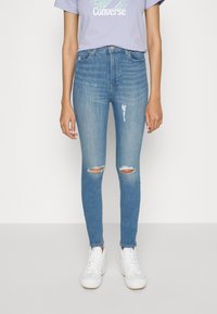 Jeans skinny azuis com rasgos nos joelhos, design de cinco bolsos e cintura média, combinados com ténis brancos e uma t-shirt lilás clara.