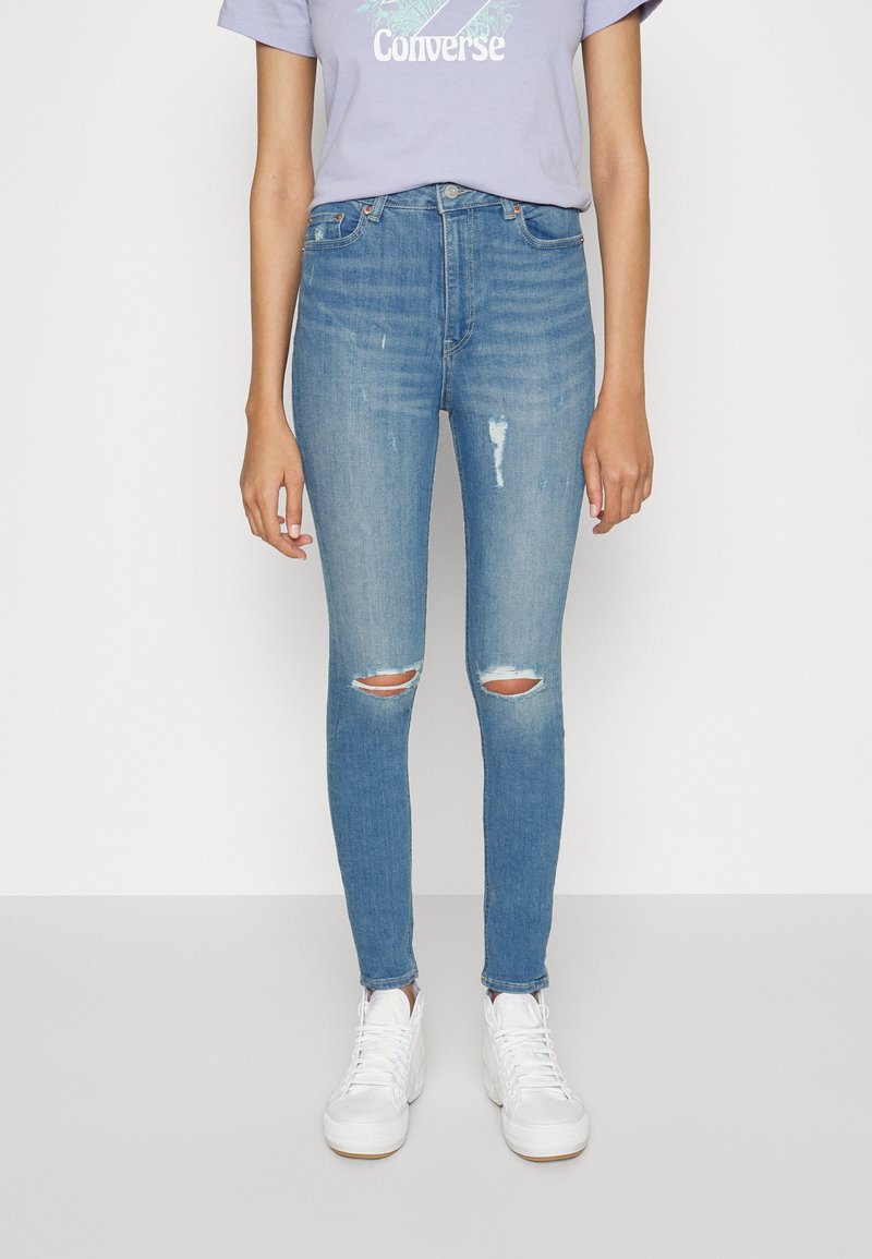 Jeans skinny azuis com rasgos nos joelhos, design de cinco bolsos e cintura média, combinados com ténis brancos e uma t-shirt lilás clara.