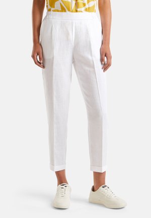 Osoba ubrana w białe, krótkie spodnie i kremowe trampki, stojąca na jednolitym tle.