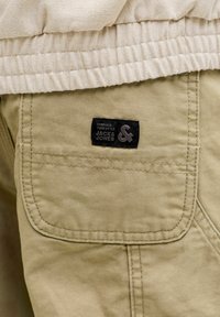 Poche d'un pantalon beige avec double couture et une étiquette en tissu noire portant l'inscription "Embrace your style JACK & JONES", assortie à un vêtement crème côtelé.