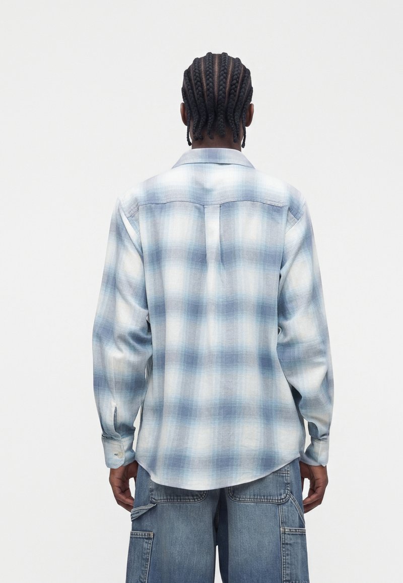 Man with braided hair wearing a blue and white plaid long-sleeve shirt and loose blue denim cargo pants, viewed from the back.