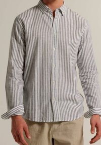 Hombre vistiendo una camisa de botones de manga larga con rayas verticales azules y blancas, combinada con pantalones beige, de pie con los brazos relajados.