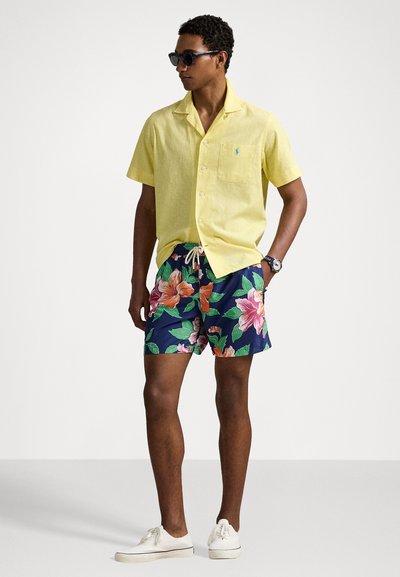 Light yellow short-sleeve shirt, navy shorts with large floral pattern, white drawstring, and white sneakers, accompanied by sunglasses.