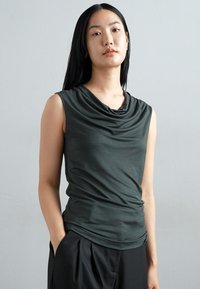Dark green sleeveless top with a cowl neck, featuring soft draping and a fitted design, paired with black high-waisted pants.