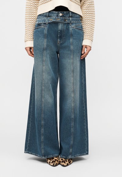 Femme portant un jean bleu taille haute à jambes larges avec des coutures devant, associé à un pull en maille beige et des chaussures pointues à imprimé léopard.