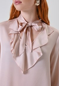 Rüschige, hellrosa Bluse mit gebundenem Ausschnitt. Weicher, glatter Stoff mit zartem Glanz und subtilen Stickdetails.