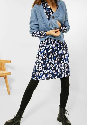 Frau, die ein blaues Leopardenmusterkleid, schwarze Strumpfhosen, schwarze Schnürstiefel und eine klobige blaue Strickjacke trägt, steht mit gespreizten Beinen und Händen, die den Pullover anpassen.