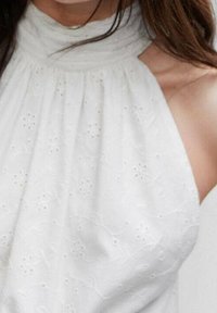 Haut halter blanc avec un décolleté festonné, orné de motifs floraux brodés et d'une finition texturée. Tissu léger qui tombe doucement.