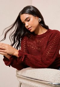 Burgundy stickad tröja med texturerade mönster och volangärmar, med band vid ärmarna. Långt hår flödar åt ena sidan.