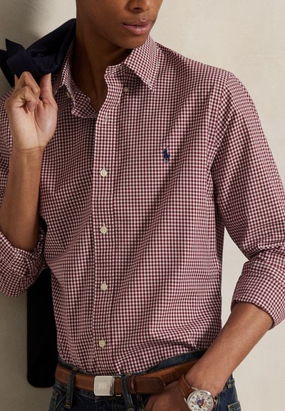 Muž nosiaci červeno-bielo kockovanú košeľu s vyhrnutými rukávmi, drží čiernu bundu cez rameno a nosí hodinky a opasok.