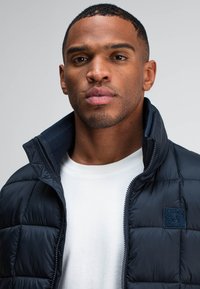 Marineblauer gesteppter Puffer-Jacke mit hohem Kragen, Reißverschluss und strukturiertem Patch-Logo. Getragen über einem weißen Rundhals-T-Shirt.