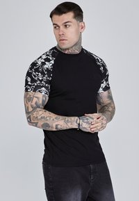 Musta lyhythihainen t-paita, jossa on mustavalkoiset kuviolliset hihat. Ominaisuuksiin kuuluu istuva muotoilu ja rintalogot, sopii arkikäyttöön.
