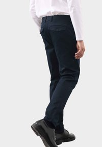 Marineblaue Hose aus glattem Stoff mit geradem Schnitt, ausgestattet mit zwei Gesäßtaschen und dezenten Markenlogos. Kombiniert mit schwarzen Schuhen.