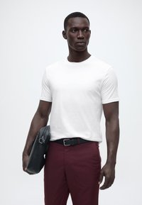 Άνδρας που φοράει λευκό κοντομάνικο πουκάμισο και μπορντό παντελόνι κρατώντας μαύρη δερμάτινη τσάντα, κοιτάζοντας προς τα πλάγια, με απλό ανοιχτόχρωμο φόντο.