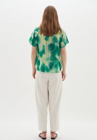 Blusa verde a maniche corte con motivo astratto, vestibilità ampia, tessuto leggero, abbinata a pantaloni plissettati crema e sandali neri.