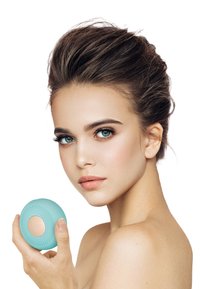 Ansiktsrenseapparat i lys blå silikon med teksturert overflate, holdt i hånden nær en models ansikt, med gullakenter.