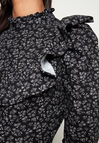 Black floral blouse featuring a high ruffled neckline, textured fabric, and puffed sleeves with ruffle accents. White floral pattern throughout.