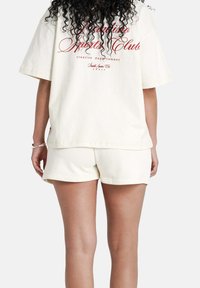 Persona con lunghi capelli ricci che indossa una maglietta oversize color crema con testo rosso e pantaloni coordinati, vista da dietro su uno sfondo semplice.