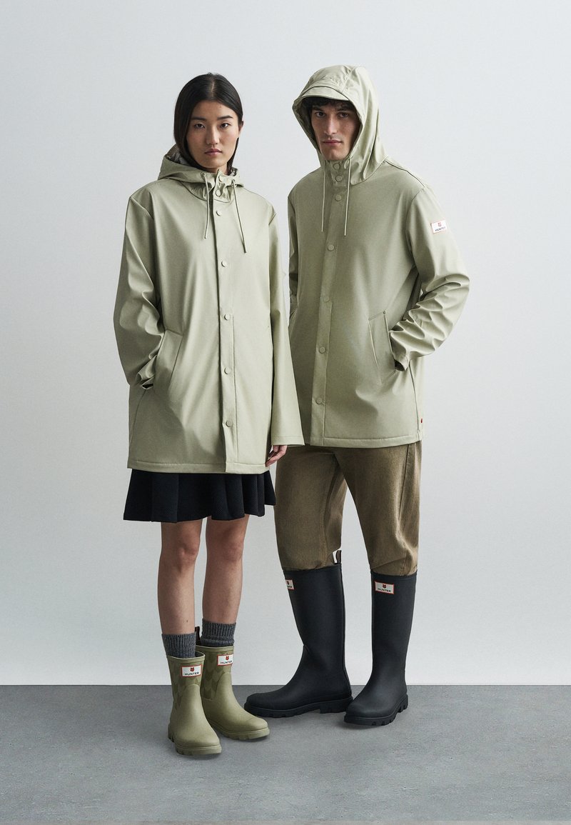 Δύο μοντέλα φορώντας ανοιχτό πράσινα αδιάβροχα και μποτάκια Hunter, στέκονται σε γκρι δάπεδο μπροστά σε λευκό απλό φόντο.