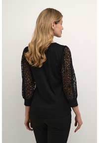 Haut noir avec des manches bouffantes en dentelle. Le corps est entièrement noir, tandis que les manches présentent un motif en dentelle floral complexe. Encolure ras du cou simple.