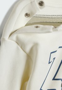 Tessuto color crema con finitura testurizzata, dotato di automatici in metallo e una stampa grafica blu. Il dettaglio ravvicinato evidenzia la qualità del materiale.