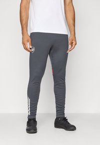 Grå atletiske bukser med slim fit, kontrasterende rød kant, hvide striber langs siderne og et broderet logo på låret. Sorte støvler iført.