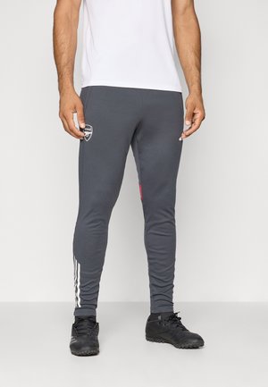Grå atletiske bukser med slim fit, kontrasterende rød kant, hvide striber langs siderne og et broderet logo på låret. Sorte støvler iført.