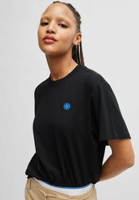 T-shirt di cotone nera con colletto rotondo e maniche corte; presenta un logo di faccina sorridente ricamato in blu sul lato sinistro del petto.