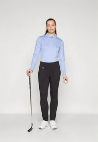 Világoskék, hosszú ujjú póló zipzáras gallérral, fekete, testhezálló nadrággal párosítva; golfütőt tart, fehér sportcipőt visel.