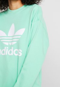 Mintgrön sweatshirt i mjukt material som har en stor vit Adidas-logotyp med tre ränder under på framsidan.